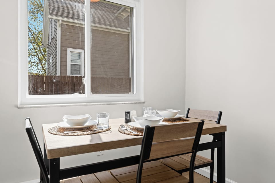 Cozy dining nook with a modern wood table set for four, bathed in natural light from a large window for an inviting mealtime atmosphere.