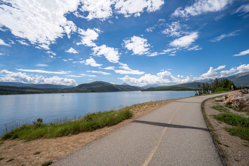 Scenic lakeside road winds through pristine mountain landscape with crystal-clear waters and forested hills under dramatic blue skies.