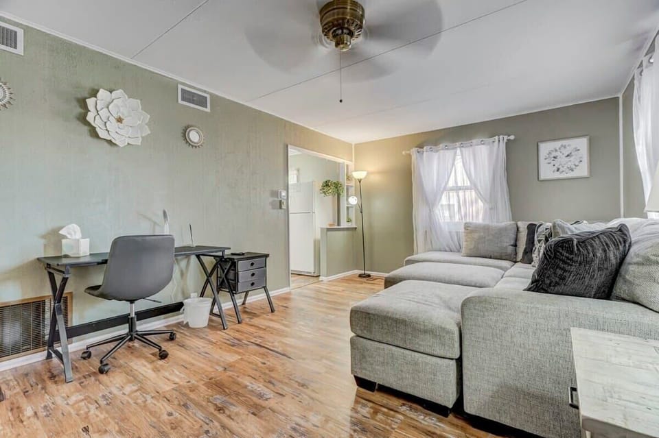 Settle in on the gray sectional under the ceiling fan, with a desk-and-chair corner nearby for planning or journaling—wood floors and airy curtains give the room a relaxed, homey feel.
