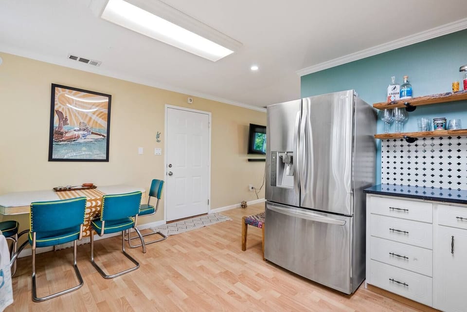 This open-concept dining area features a retro-style table with vibrant teal chairs, set against light neutral walls and wood-toned flooring. The space flows into a modern kitchen with a large stainless steel refrigerator.