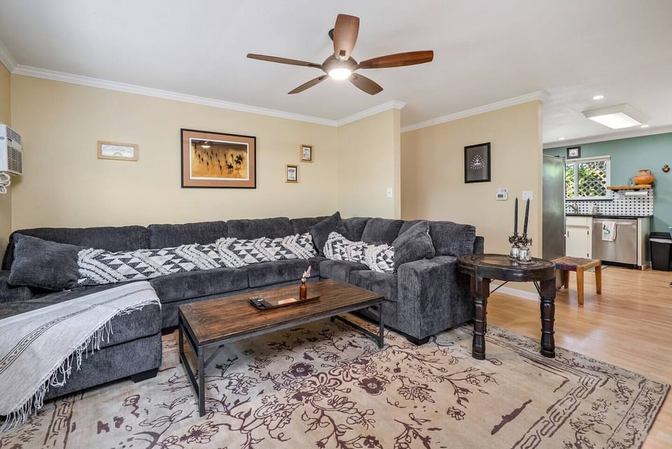 This open-concept living space features a massive grey L-shaped sectional with geometric pillows, a rustic wood coffee table, and an ornate floral rug. The room flows seamlessly into the kitchen, creating a perfect setup for relaxing or entertaining.