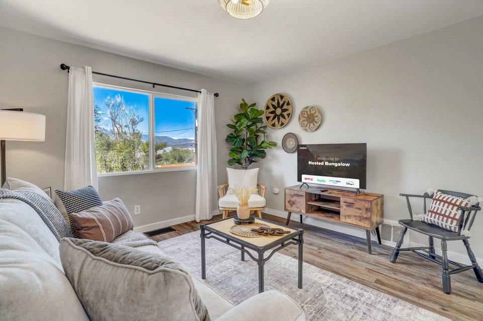 Welcome to our inviting living room, where comfort meets style. Two cozy chairs by the TV, an elegant sofa adorned with plush cushions, and a tasteful coffee table with a vase create a charming ambiance. Lush tree views on the left, an exquisite frame above the sofa, and soft, inviting lighting complete the scene. An accessible outlet is also provided for your convenience.