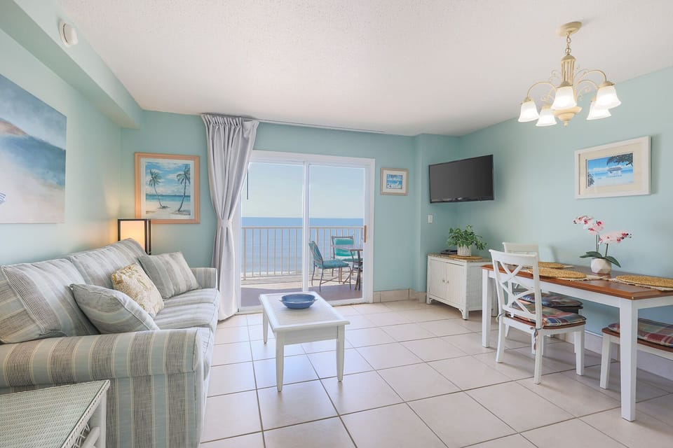 This inviting living area features a light blue color scheme, a cozy sofa, a TV, and a stunning beach view from the balcony