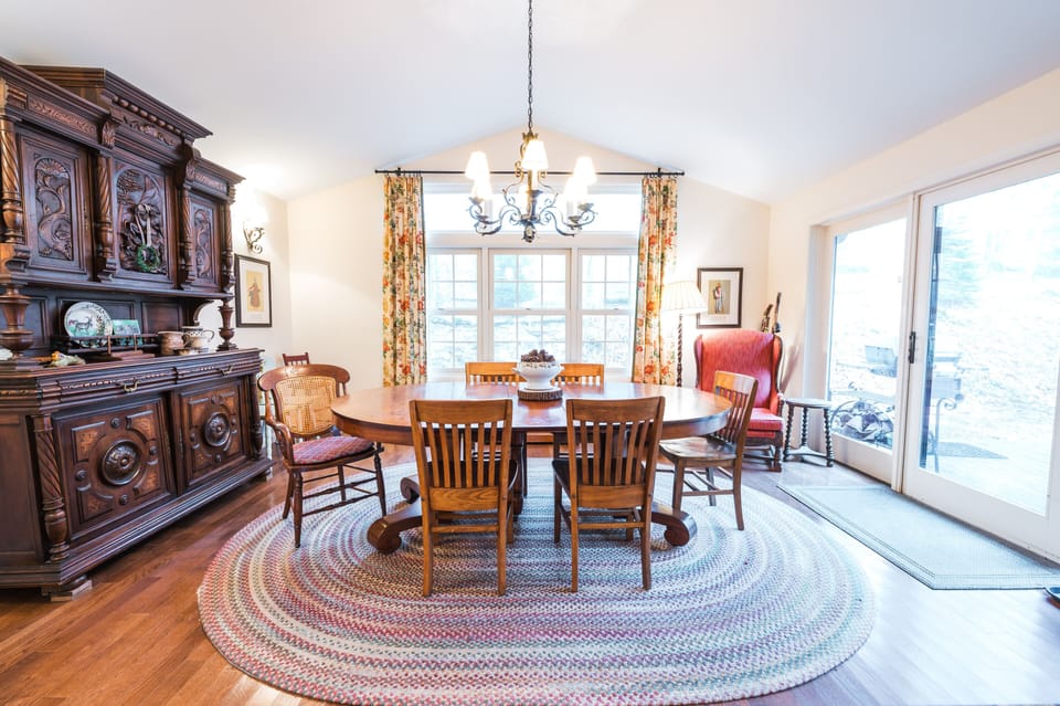 Wooden dining set with antique hutch, large windows, and floral curtains.