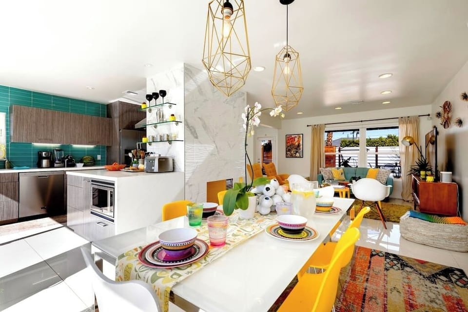 Bright open-plan dining and kitchen with gold geometric pendants, teal tile backsplash, and bold colorful seating — where the group gathers after a pool day