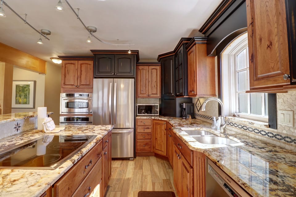 A modern kitchen featuring a stainless steel refrigerator, granite countertops, wooden cabinets, an electric stovetop, a dishwasher, and a window above the sink. Track lighting illuminates the space.