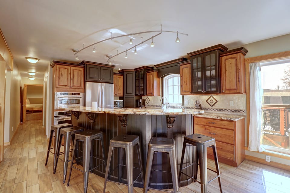 A modern kitchen with a curved island, surrounded by six metal barstools. It features mixed wood cabinets, stainless steel appliances, and a tiled backsplash. A sliding glass door leads outside.