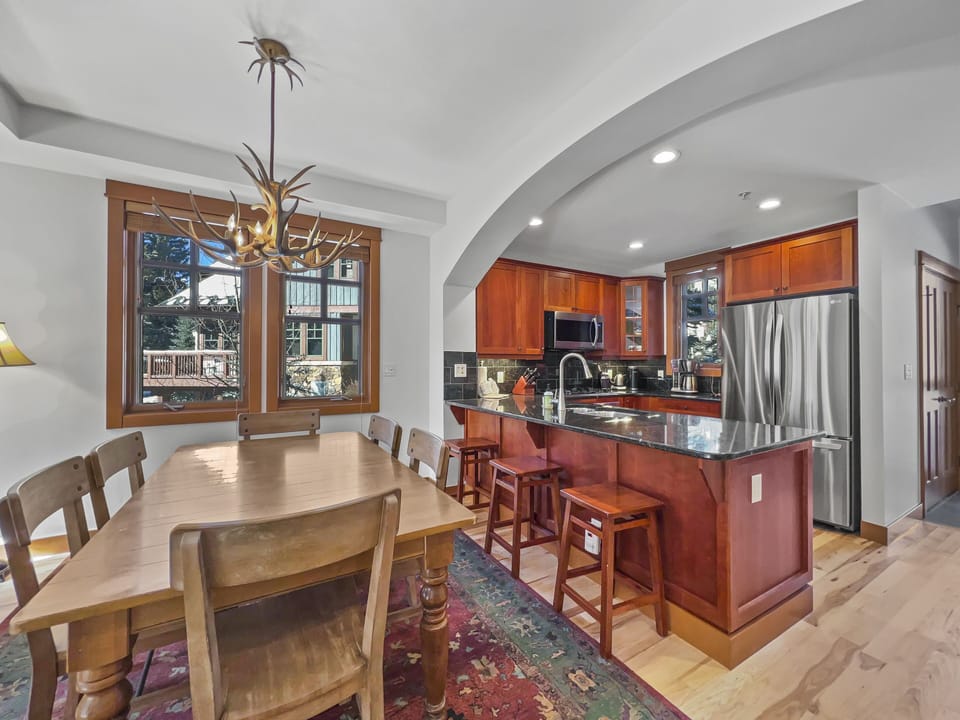 A cozy kitchen and dining area with wooden furniture, a wooden dining table, stainless steel appliances, and a modern island with bar stools. A large window provides natural light.