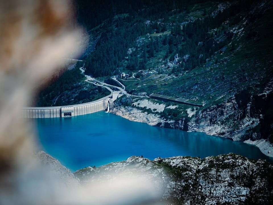 An aerial view of a vivid blue reservoir nestled between steep, snow-dusted mountain cliffs.