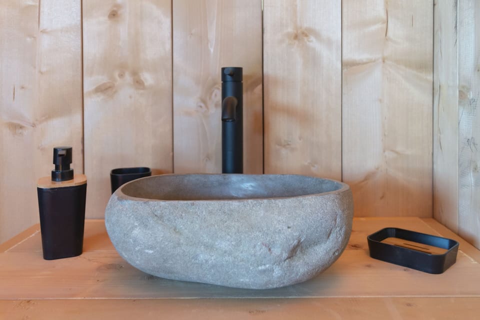 Exclusive natural stone basin as the centerpiece of a refined luxury bathroom. Clean lines and matte black fixtures create a striking contrast with warm wooden walls, enhancing the sense of understated elegance. Thoughtful details elevate the experience, delivering high-end wilderness comfort immersed in nature.