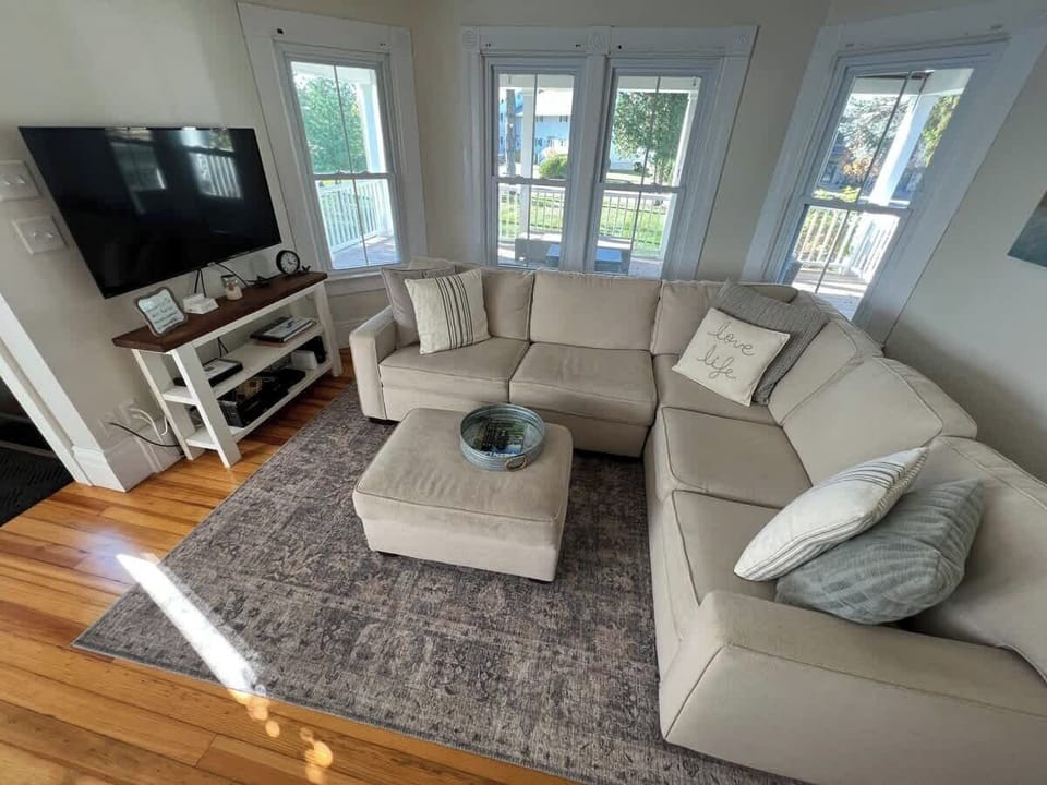 The cozy Living Room is furnished with a large sectional and TV. The bay window provides natural lighting with views out to the Lake across the street.