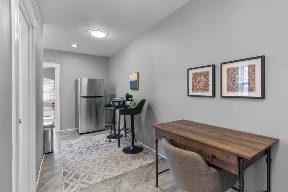 A dedicated wood desk with upholstered office chair shares the tiled hall with a stainless refrigerator, round high table, two green stools, patterned runner, and doorway toward the living room.