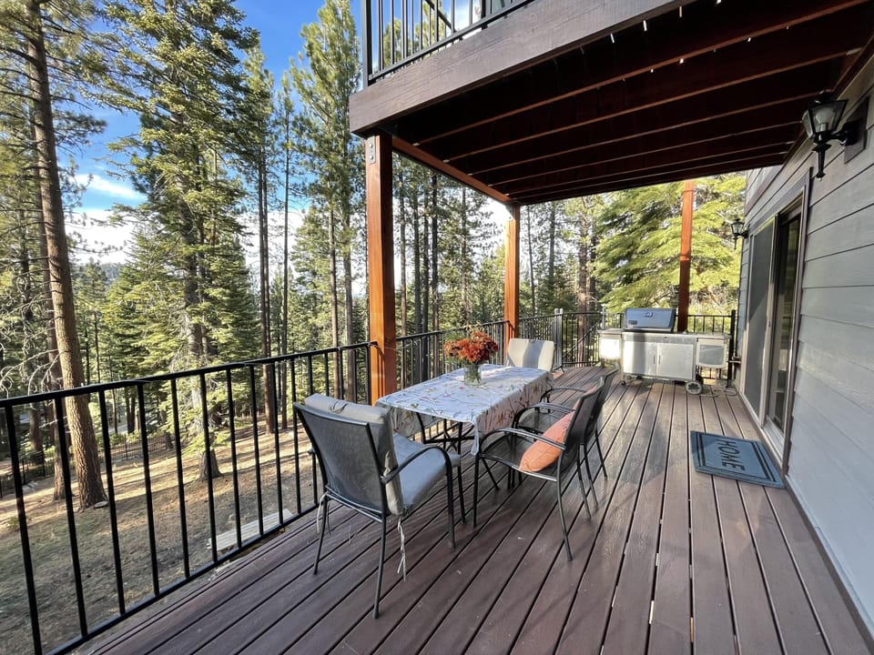 Covered wooden deck with black metal railing, outdoor dining table, cushioned chairs, and stainless steel grill.