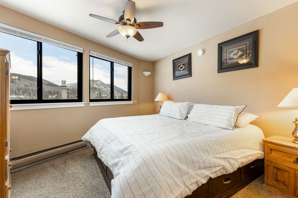 Bedroom with a large bed, white-striped bedding, two nightstands with lamps, framed pictures on beige walls, three windows with mountain views, and a ceiling fan.