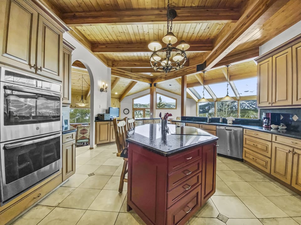 Spacious kitchen with wooden ceiling, island, stainless steel appliances, double ovens, and scenic windows. Cream cabinetry and tiled floor contrast with a dark granite island countertop.