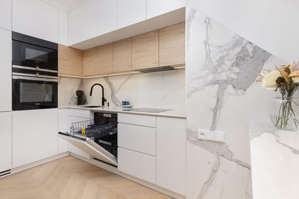 A modern kitchen with white cabinets and wooden accents, including built-in appliances and a glass door leading to the balcony.