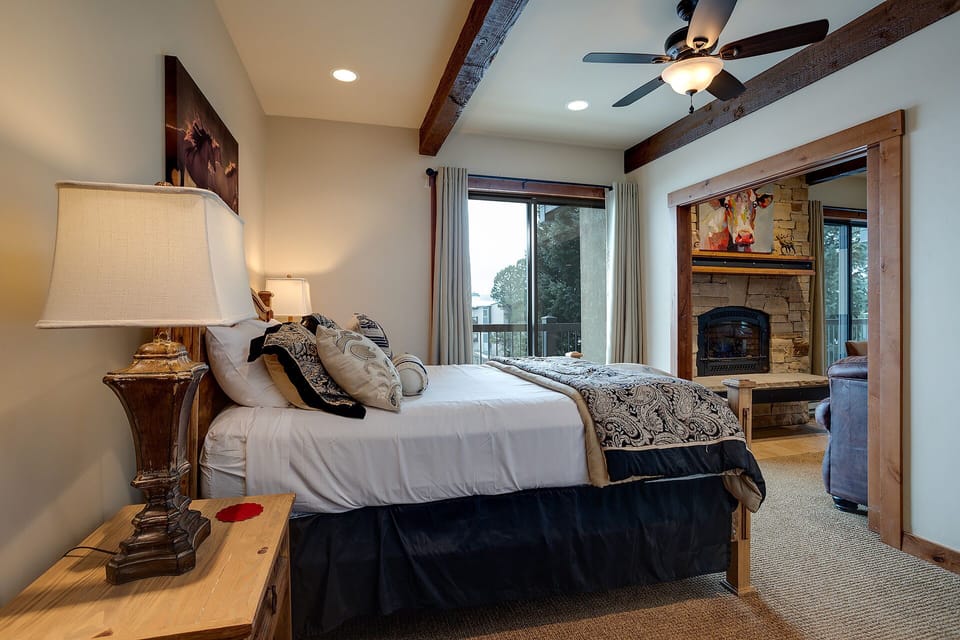 Unwind in your cozy bedroom featuring exposed beams, a stone fireplace, and serene views—perfect for peaceful mountain mornings.