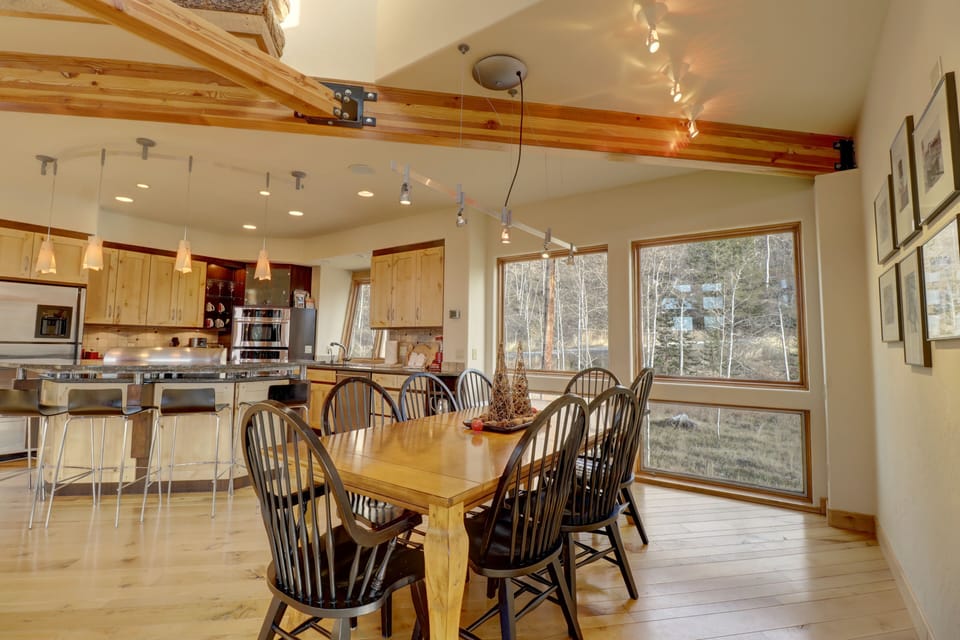 A modern kitchen and dining area with a wooden table and black chairs. The kitchen features wooden cabinets, stainless steel appliances, and pendant lighting. Large windows provide natural light.