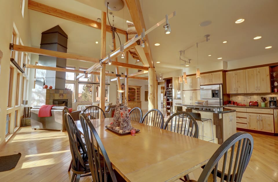 A spacious kitchen and dining area with wood furnishings, high ceilings, and ample natural light from large windows. The dining table is set for eight, and there's a modern kitchen island and appliances.