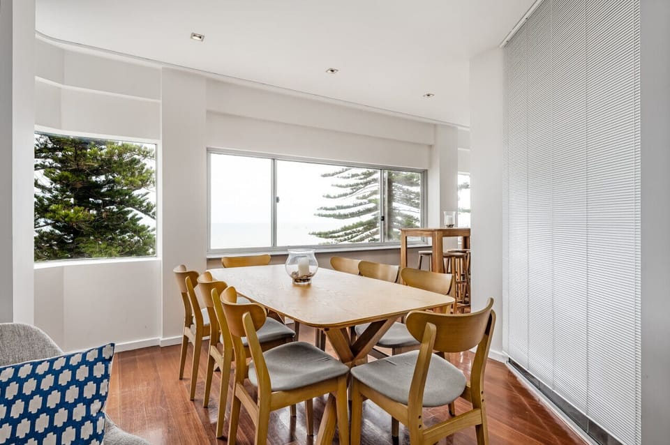 This charming dining area seamlessly blends natural warmth with a clean, minimalist aesthetic. At the center stands a beautifully crafted wooden dining table, its surface smooth and slightly textured to showcase the rich grain and organic imperfections of