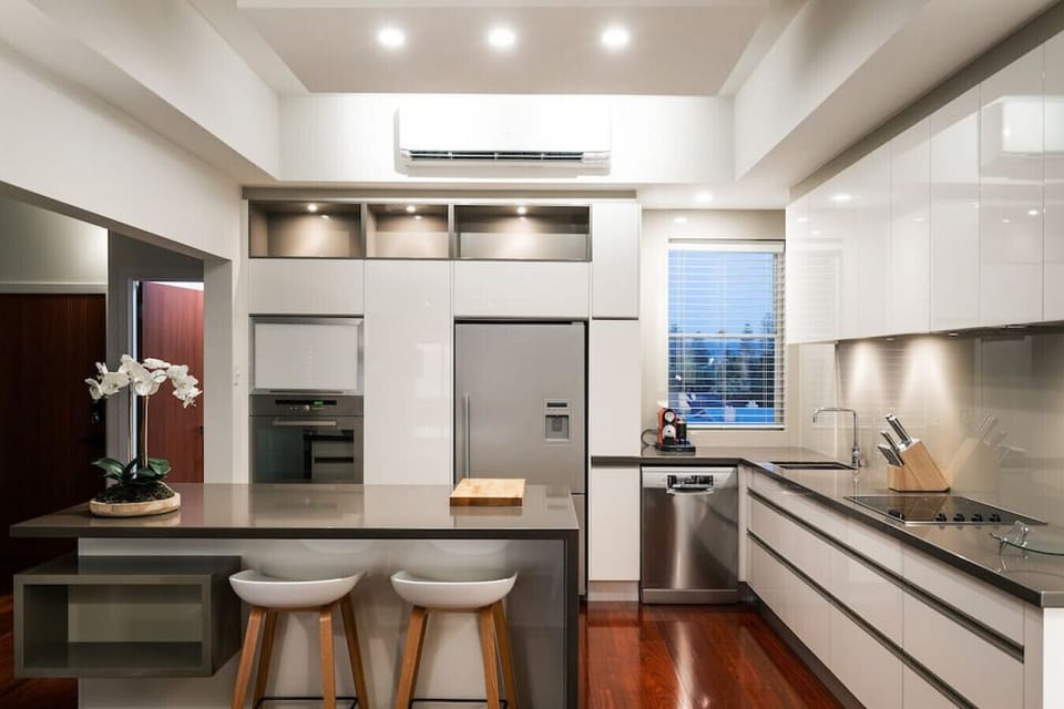 This modern kitchen features sleek white cabinetry, stainless steel appliances, and a glossy island with bar seating. Under-cabinet lighting and rich wood floors add warmth to the clean, contemporary design.