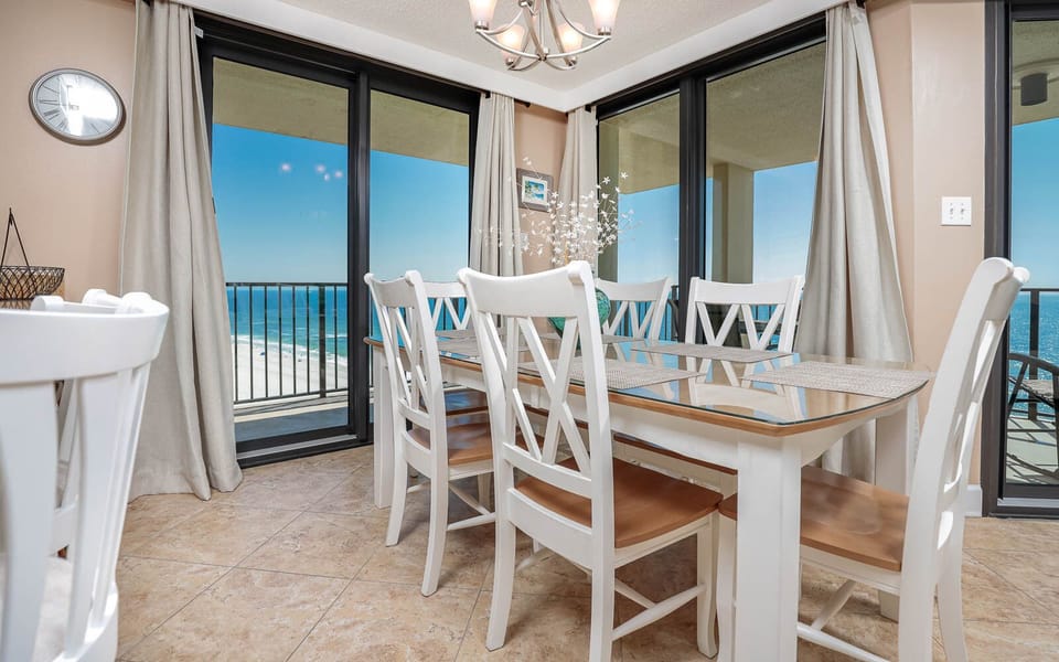 A bright dining area features a glass-top table with white chairs and offers a stunning view of the beach