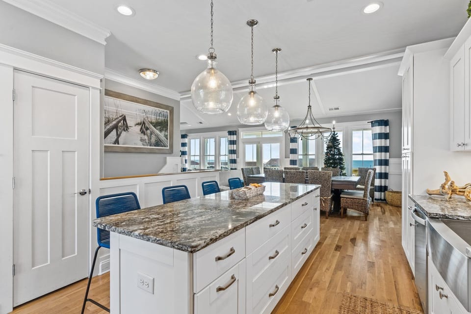 Bright coastal kitchen with white cabinetry, granite island, blue barstools, stainless appliances, pendant lighting, and deck access.