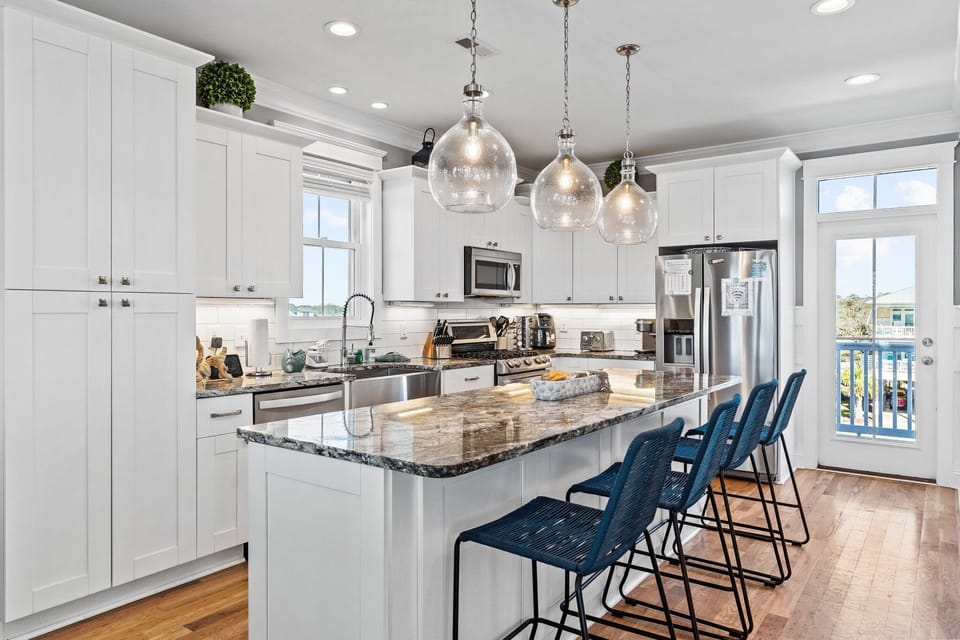 Bright coastal kitchen with white cabinetry, granite island, blue barstools, stainless appliances, pendant lighting, and deck access.