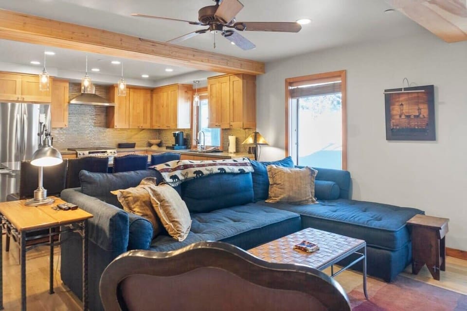 The first living room features a large sectional sofa with a chaise on the right side, a leather chair, a mounted 37” flat-screen TV, and a stone hearth double-sided fireplace.