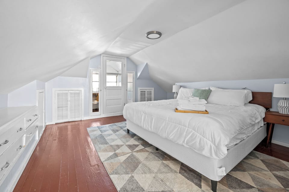 This attic bedroom angle includes a white-dressed bed, built-in drawer storage, bedside lamps, sloped ceilings, wood floors, geometric rug, and doorway toward the windowed work nook.