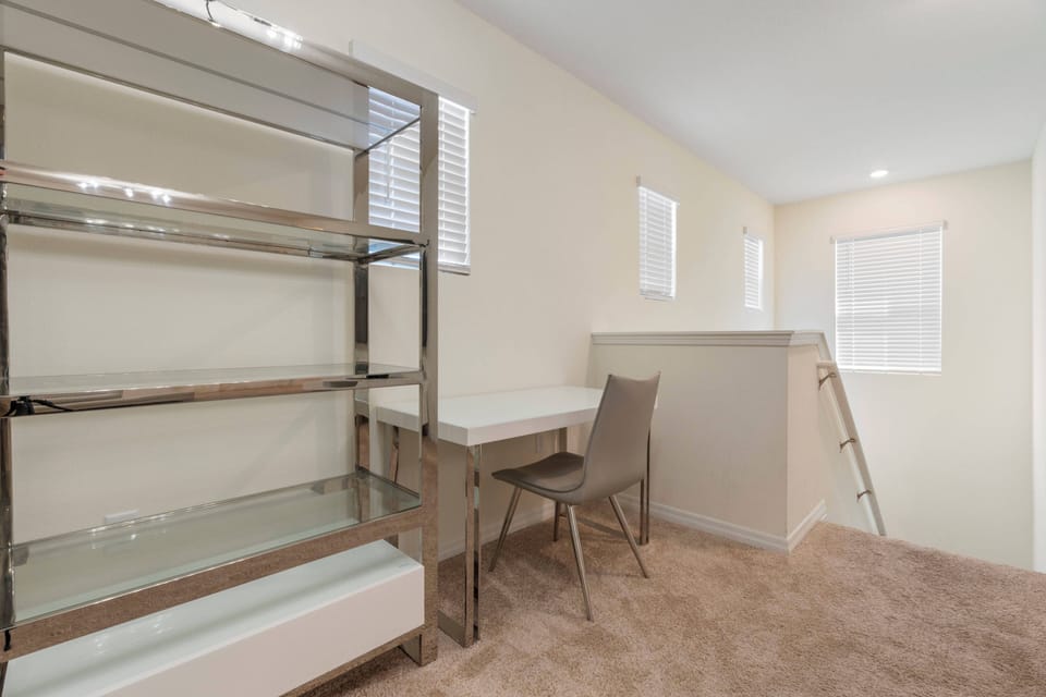 This bright and airy room offers a sleek glass shelving unit and a minimalist desk with a modern chair, ideal for relaxing or working. Large windows let in natural light, creating an inviting atmosphere for your stay.