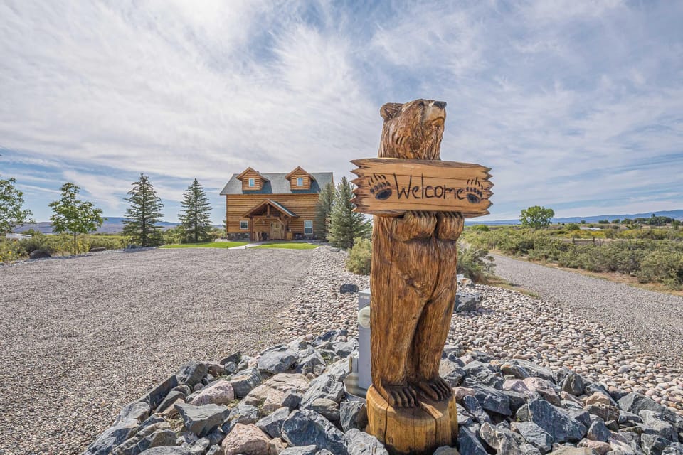 Hand-carved wooden bear stands on river rocks, holding a welcome sign at the gravel driveway entrance.