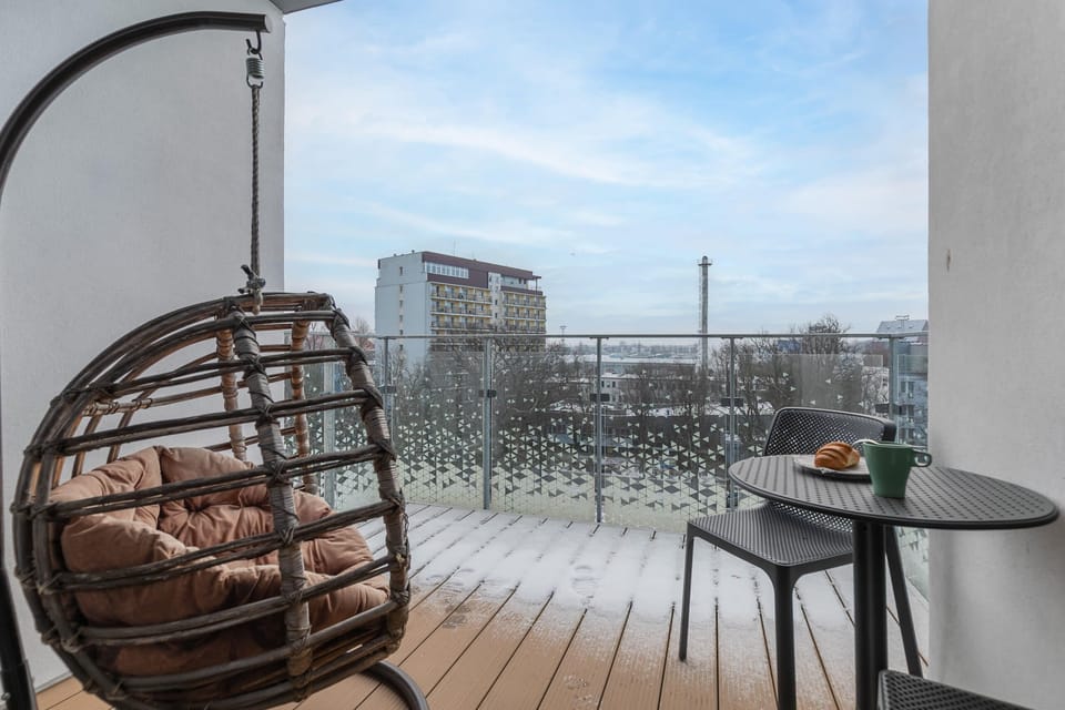 A balcony with a hanging wicker chair and a small table, offering scenic city views.
