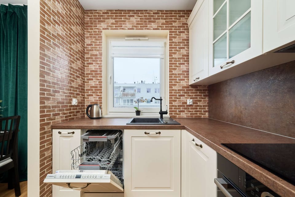 A kitchen with white cabinets, dark wooden countertops, and a brick accent wall, with a kettle and orchid on the counter.