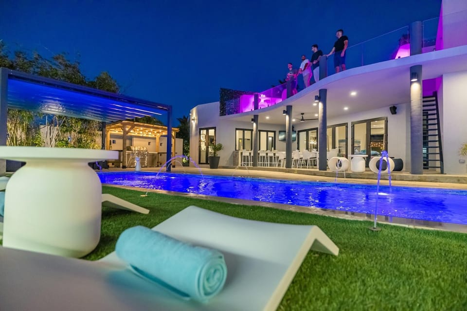 Luxurious villa with a long pool, white lounge chairs with blue towels, palm trees, and modern white architecture creating a bright, tropical retreat.