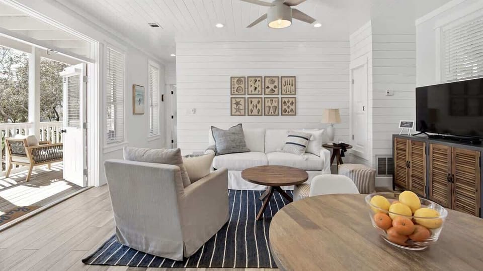- Bright coastal living room with a plush white sofa & accent chair for lounging in style - Nautical wall art & a bold striped rug add texture and beach inspired flair - Light wood tones, TV setup, and soft hues create a relaxing retreat indoors