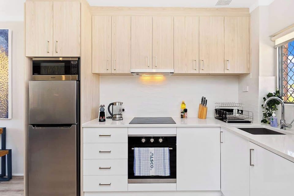 This modern kitchen features a refrigerator, ample cabinets, stove, sink with faucet, Nespresso, electric kettle, and a salt & knife set, all bathed in light from the kitchen window. 