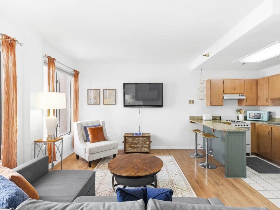 The stylish living room boasts a large sectional couch, a cozy couch chair, a coffee table, a stylish rug, a mounted 50” flatscreen TV, two side tables, and lamps offering the perfect amount of lighting for a cozy ambiance.