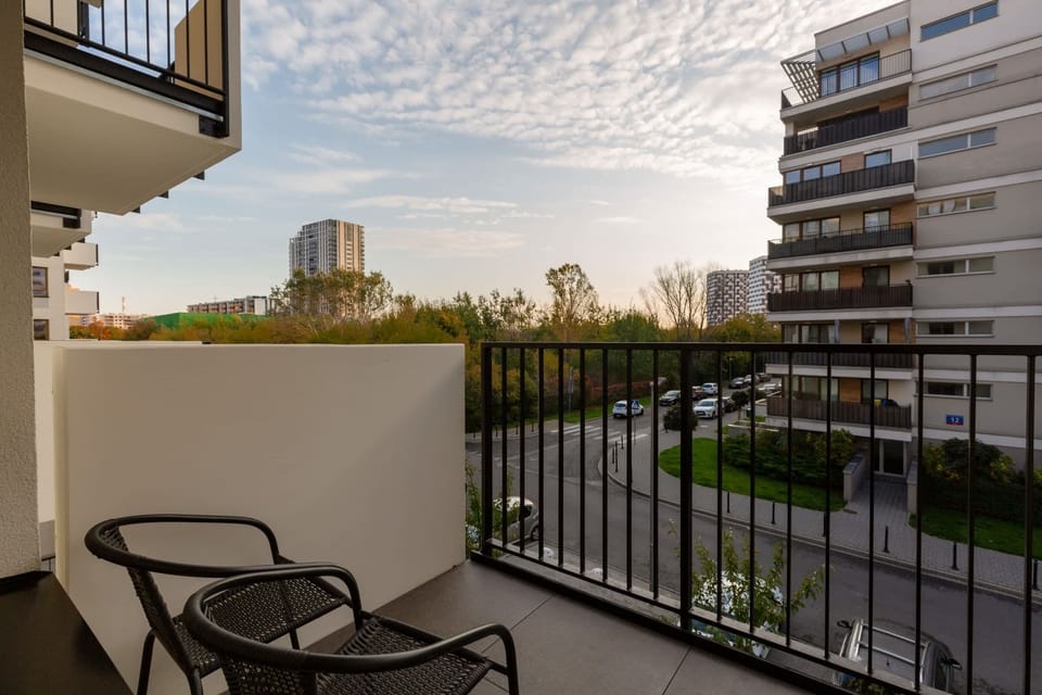 A view of the balcony, furnished with chairs and a table. Greenery and buildings can be seen in the distance.
