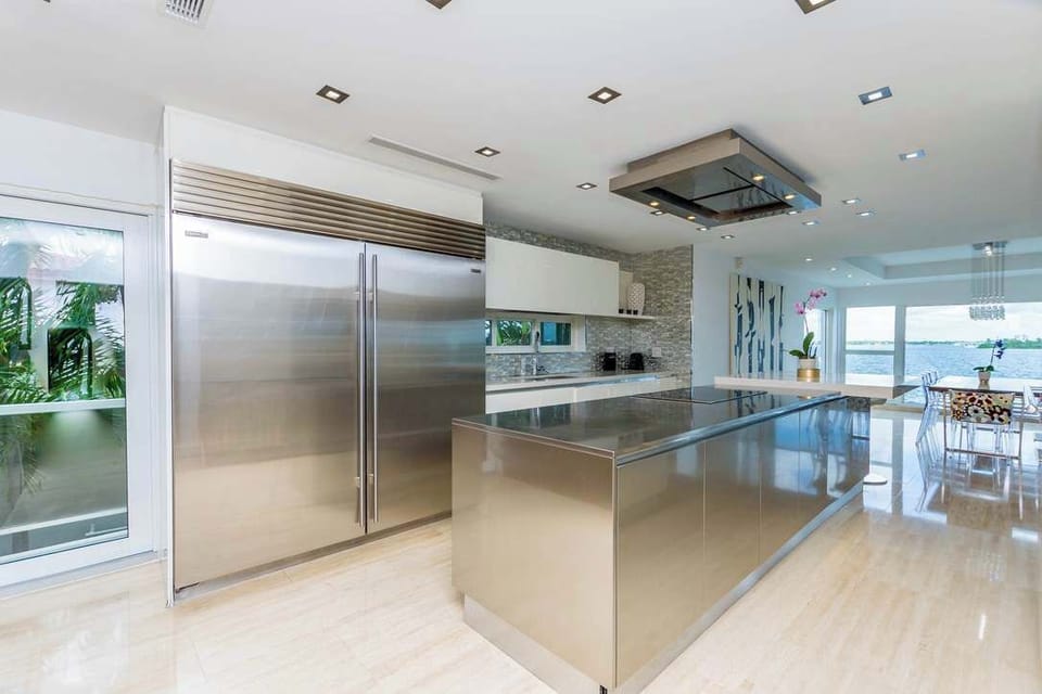 Chef’s kitchen featuring a large stainless steel island, commercial-grade refrigerator, built-in cooktop with ceiling-mounted range hood, custom cabinetry, recessed lighting, and an open-concept layout connected to the dining and living areas.