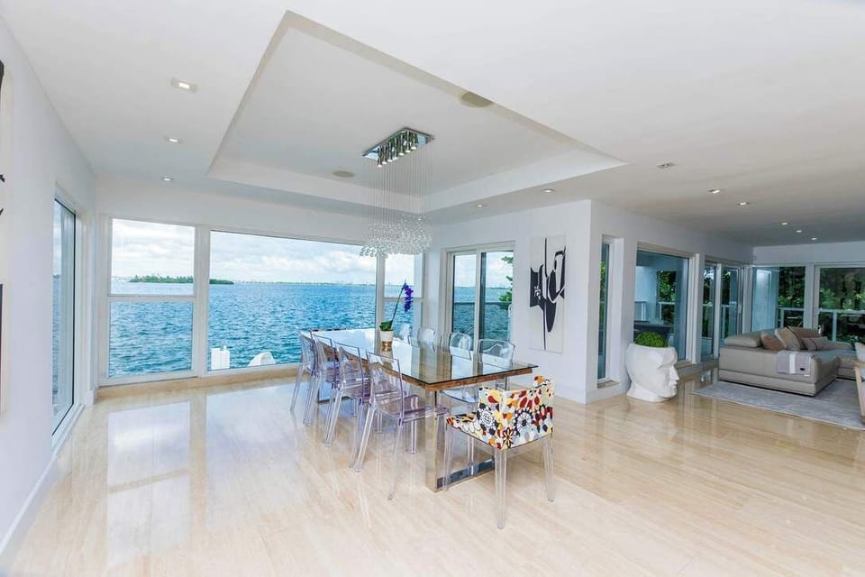 Waterfront dining area featuring a glass dining table with seating for eight, floor-to-ceiling windows, panoramic bay views, recessed lighting, tile flooring, and an open-concept layout connecting the living room, terrace, and entertaining spaces.