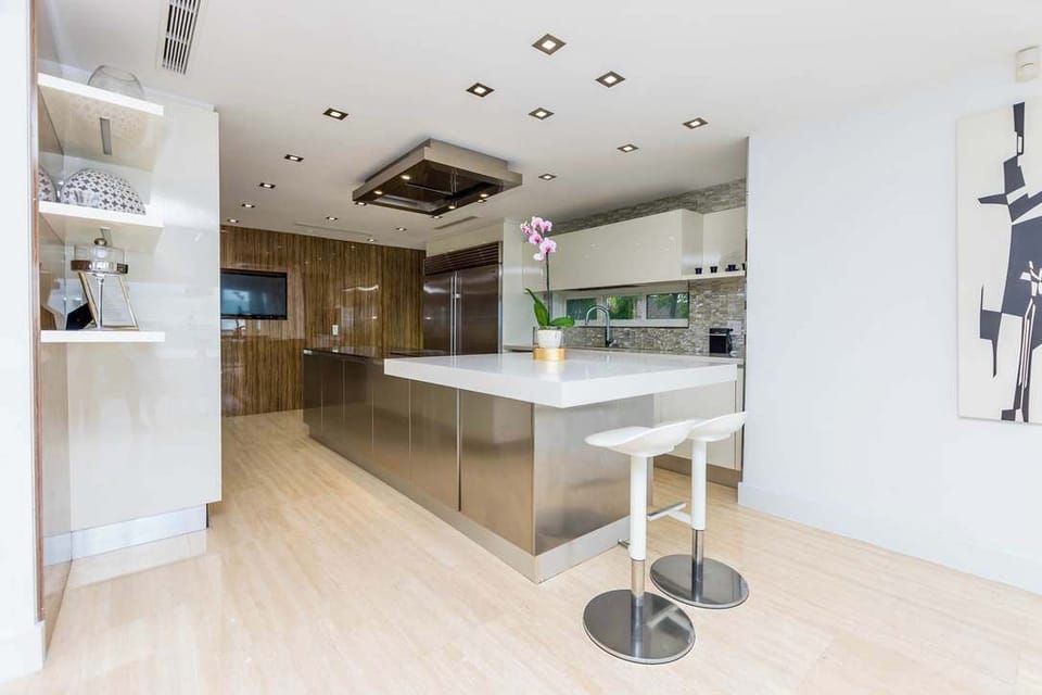 Contemporary kitchen workspace featuring a large waterfall-edge island, stainless steel cabinetry, breakfast seating, custom wood-paneled storage, recessed lighting, built-in appliances, and an open layout connected to the dining and living areas.