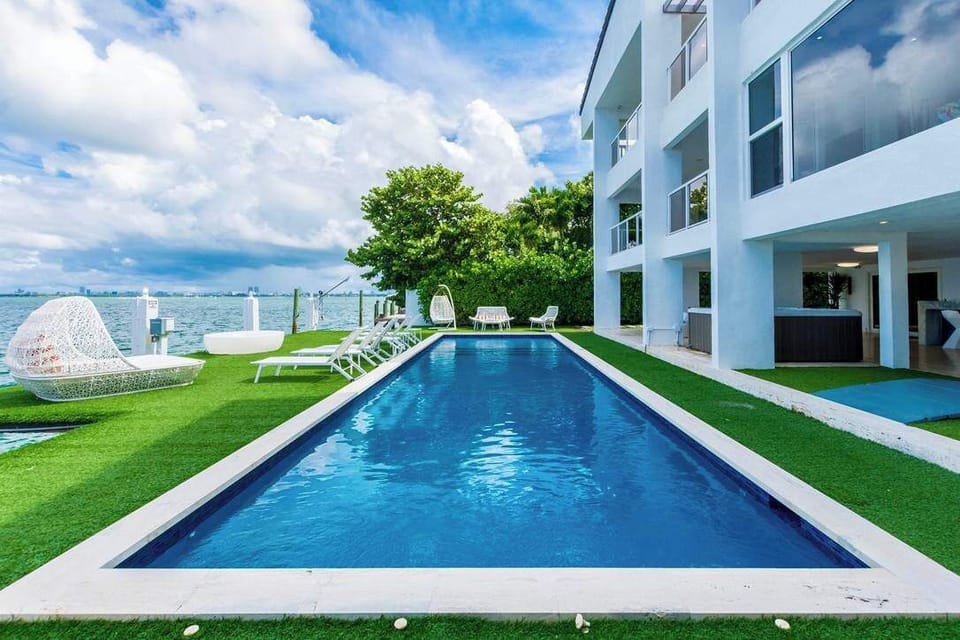 Waterfront outdoor area featuring a private lap pool, artificial turf lawn, poolside seating, glass safety fencing, covered patio space, and panoramic water views. Located alongside a modern multi-story villa with direct access to outdoor amenities.