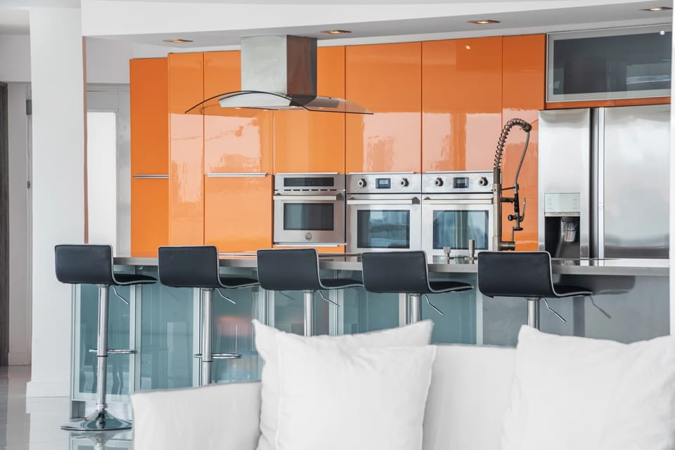 Modern kitchen featuring glossy orange cabinetry, stainless steel appliances, double wall ovens, French-door refrigerator, glass breakfast bar with seating for six, built-in microwave, and open-concept layout overlooking the living area.