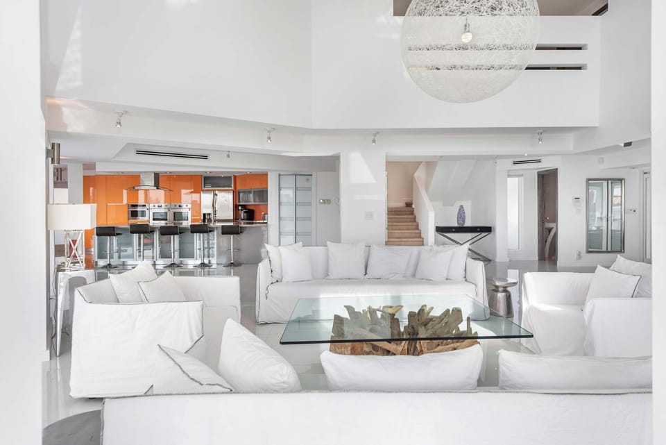 Bright open-concept living space with white sectional seating, glass coffee table, designer chandelier, vaulted ceilings, glossy tile floors, modern kitchen with bar seating, and convenient access to upper-level bedrooms and common areas.