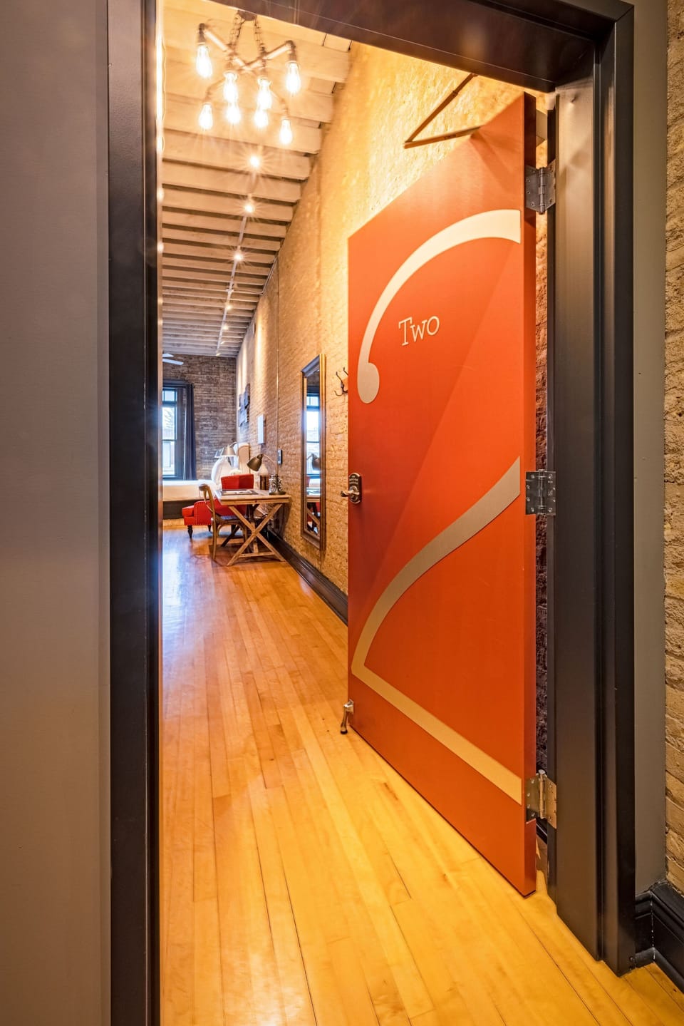 The corridor opens into the full length of the loft — exposed timber ceiling, brick wall, warm hardwood floors.