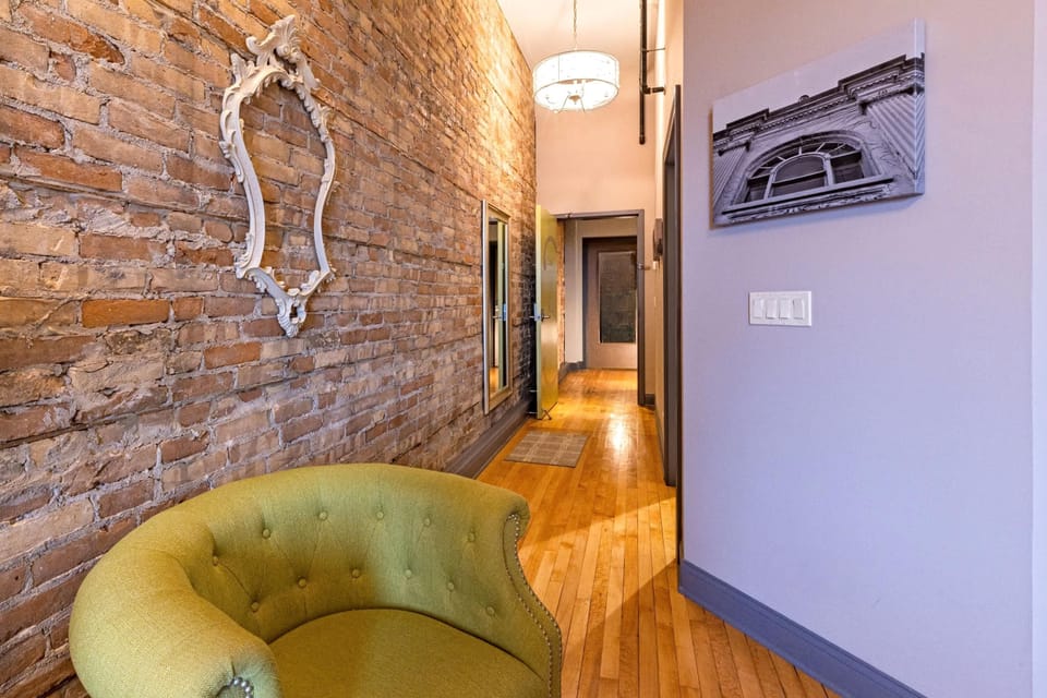 A tufted green chair, ornate mirror on brick, and a long hardwood hallway leading to your private entrance. The whole loft in one glance.