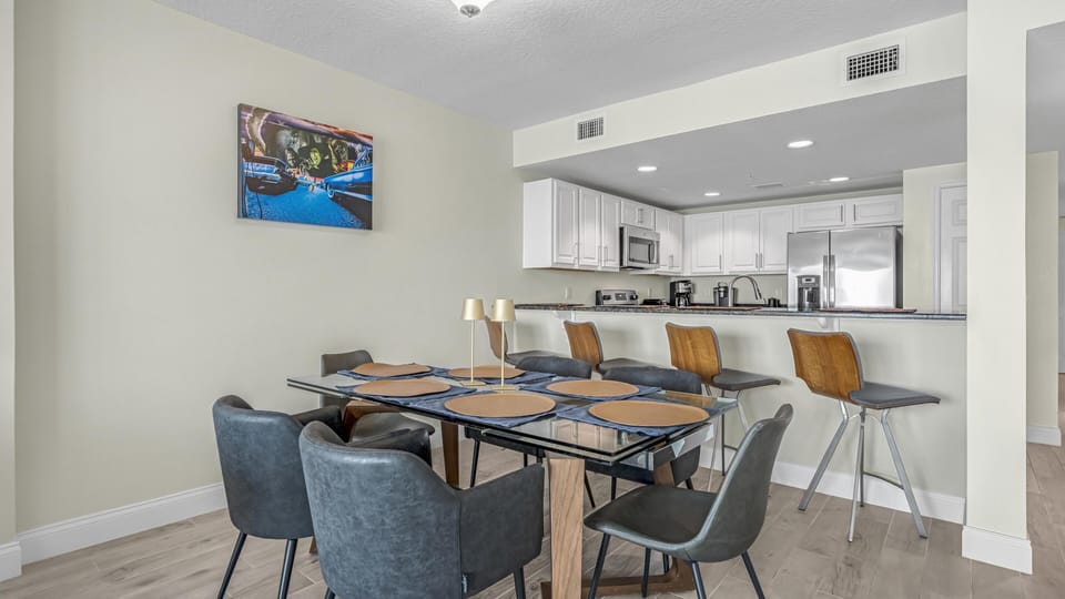 Glass dining table with gray leather chairs and tan placemats sits adjacent to the kitchen bar with wood-back stools.
