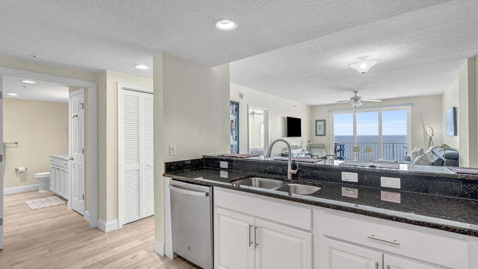 Black granite countertop with double sink overlooks living room and ocean through sliding glass doors.
