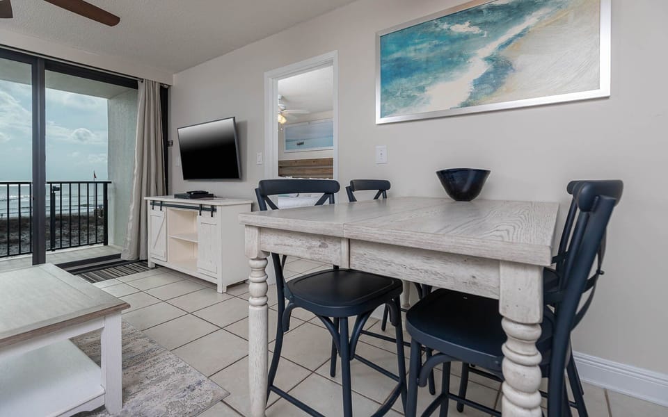 The living area features a large TV, a cozy dining table, and a stunning beachfront view through sliding glass doors
